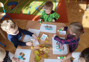 Dzieci przyklejają wycięte z papieru kolce jeża na kolorowance, następnie wyklejają wokół jeża liście