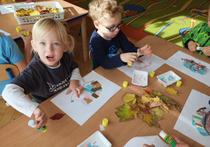 Dzieci przyklejają wycięte z papieru kolce jeża na kolorowance, następnie wyklejają wokół jeża liście