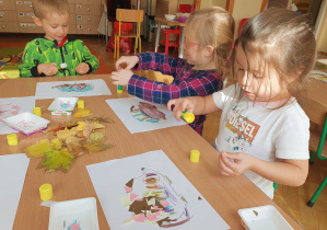 Dzieci przyklejają wycięte z papieru kolce jeża na kolorowance, następnie wyklejają wokół jeża liście