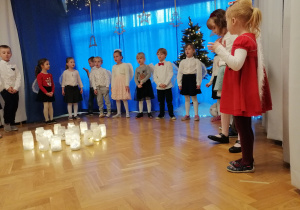 Dzieci stoją w półkolu wokół lampionów i prezentują piosenkę