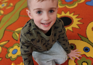 Chłopiec siedzi na dywanie i uśmiecha się do zdjęcia.