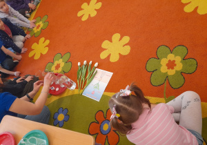 Doświadczenie - barwienie tulipanów. Dzieci siedzą na dywanie i patrzą na nauczyciela