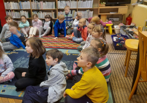 Dzieci siedzą na dywanie i biorą udział w zajęciach bibliotecznych