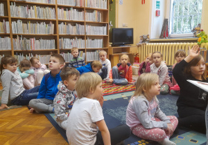 Dzieci siedzą na dywanie i biorą udział w zajęciach bibliotecznych