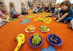 Dzieci siedzą na dywanie i utrwalają nazwy owoców.