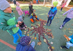 Dzieci układają kształt drzewa na boisku przedszkolnym a następnie wypełniają go kasztanami i uzupełniają liśćmi.