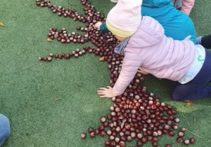 Dzieci układają kształt drzewa na boisku przedszkolnym a następnie wypełniają go kasztanami i uzupełniają liśćmi.