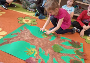 Na zdjęciu dzieci doklejają liście do wcześniej namalowanego drzewa.