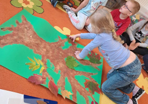Na zdjęciu dzieci doklejają liście do wcześniej namalowanego drzewa.