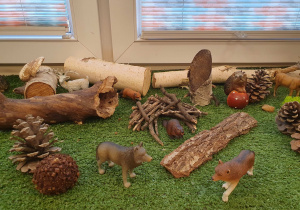 Zdjęcie przedstawia figurki wilków w otoczeniu materiału przyrodniczego takiego jak szyszki, kora drzew, pieńki, gałązki