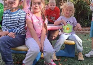 Na zdjęciu uśmiechnięte dzieci siedzą sobie w ogrodzie przedszkolnym na ławeczce.