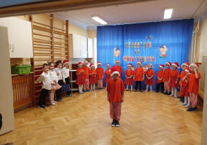 Zdjęcie przedstawia ustawione w półkolu dzieci biorące udział w przedstawieniu. Na środku chłopiec recytuje wiersz.