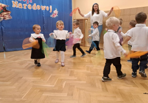 Dzieci na przedszkolnej scenie prezentują układ taneczny z kolorowymi chustami.